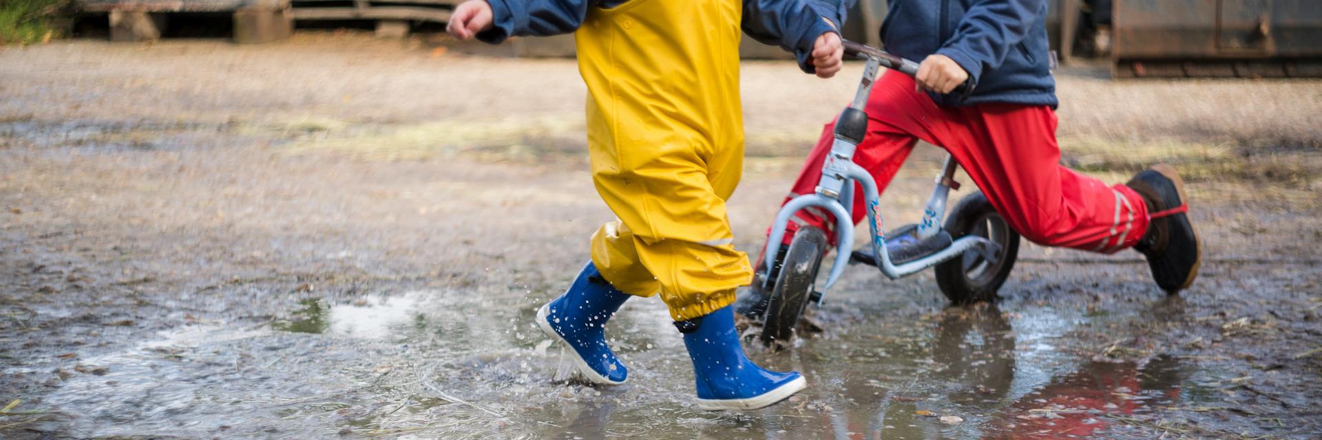 Spielende Kindergartenkinder in Pfütze mit Roller und Gummistiefel
